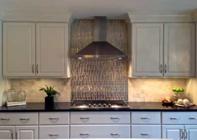 Kitchen design featuring white shaker cabinets, a dark countertop, stainless steel range hood, and a mosaic tile backsplash with warm under-cabinet lighting. - Alpharetta GA by Simone Feldman Designs