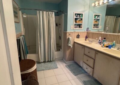 Light blue bathroom with white fixtures, striped shower curtain, dual sinks, toiletries, wicker hampers, and wooden cabinets.