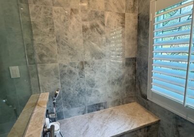 Luxury bathroom shower with marble tile walls, bench seating, handheld shower head, geometric floor tiles, and shuttered window.