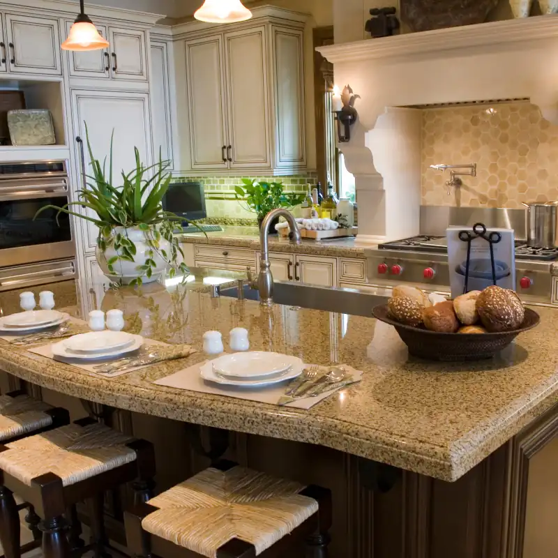 Luxury beige kitchen with granite island, stools, place settings, potted plants, and stainless steel appliances, designed by Simone.
