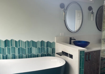 Modern bathroom design with teal hexagonal tile accents, deep teal freestanding bathtub, oval mirrors, and blue vessel sink.