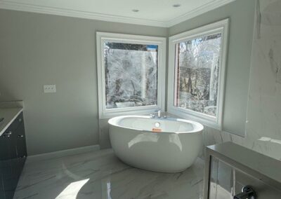 Luxury bathroom with freestanding white tub, gray walls, marble flooring, large corner windows, and modern black cabinetry.